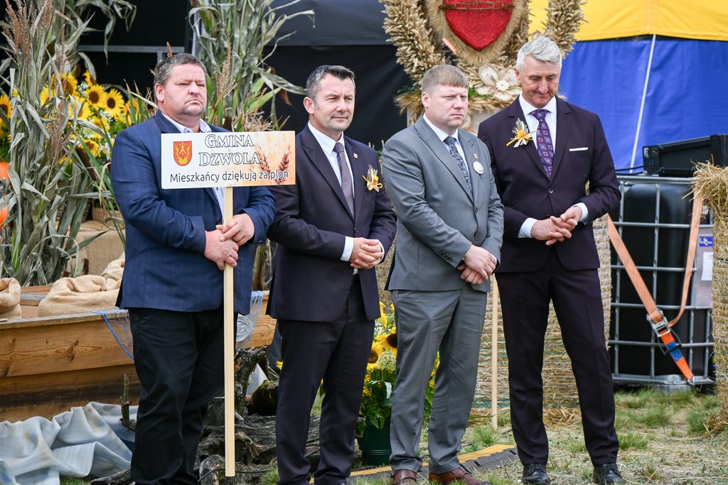 Czterech mężczyzn w garniturach, jeden trzyma tablicę z napisem „Gmina Dzwola Mieszkańcy dziękują za plon”, w tle dekoracje z kłosów zbóż i słoneczników.