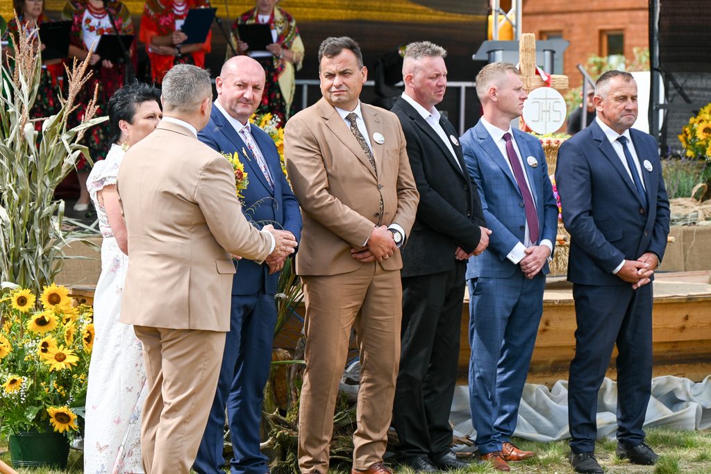 Grupa sześciu mężczyzn w garniturach i kobieta w białej sukni stoją na tle dekoracji z słoneczników i kłosów zbóż, za nimi kobiety w kolorowych strojach ludowych.