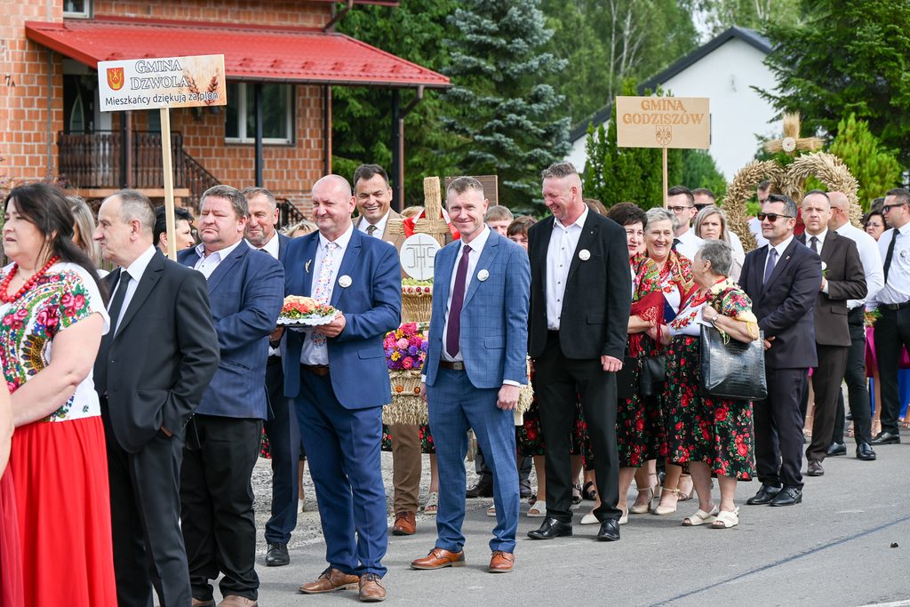 Grupa ludzi w strojach formalnych i ludowych stoi na ulicy, dwie osoby trzymają tablice z nazwami gmin, w tle zieleń i budynki.