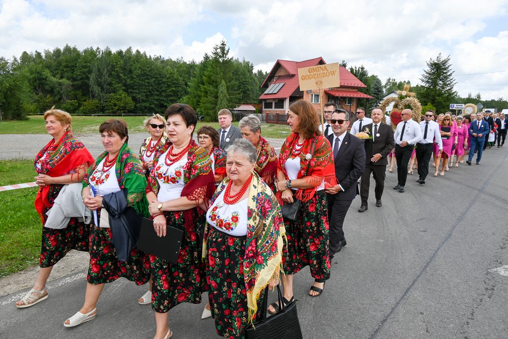 Grupa kobiet w tradycyjnych strojach ludowych z czerwonymi i zielonymi chustami, koralami, mężczyźni w garniturach idący w pochodzie na asfaltowej drodze, w tle dom i las.