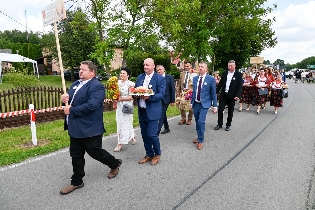 Grupa ludzi w eleganckich strojach maszeruje ulicą, mężczyzna z przodu trzyma tabliczkę, kobiety w kwiecistych sukienkach idą w rzędzie, zielone drzewa i domy w tle.