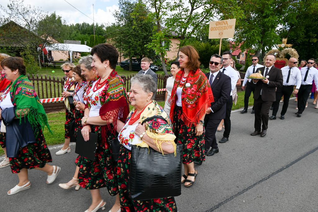 Grupa osób w tradycyjnych strojach ludowych z kolorowymi chustami i koralami idzie drogą, mężczyzna niesie bochen chleba, w tle tablica z napisem "Gmina Godziszów".
