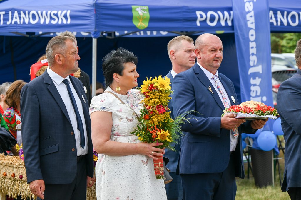 Cztery osoby w eleganckich ubraniach, kobieta trzyma bukiet kwiatów, mężczyzna trzyma tacę z chlebem, w tle niebieskie namioty i flagi z białym napisem.