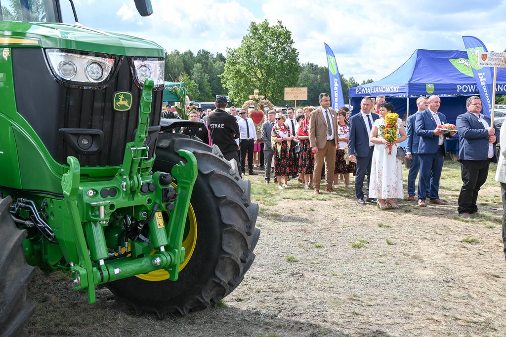 Zielony traktor obok grupy ludzi w kamizelkach i garniturach, kobiety w sukienkach z kwiatami, niebieskie namioty i flagi z napisem Powiat Janowski, znak Straż na mundurze.