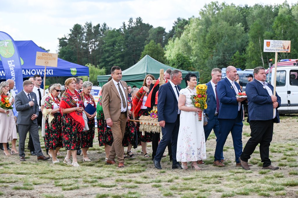 Grupa ludzi w strojach ludowych i garniturach na otwartym terenie, kobiety w kwiecistych spódnicach i czerwonych chustach, mężczyźni trzymający tablice z napisami, namioty i drzewa w tle.
