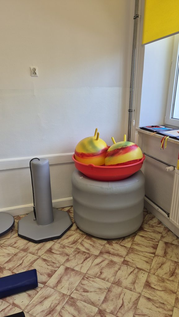 Dwa żółte piłki do skakania z czerwonymi i zielonymi smugami w czerwonej plastikowej misce na dużym szarym podwyższeniu, obok stoi szare urządzenie na podłodze z brązowymi kaflami.