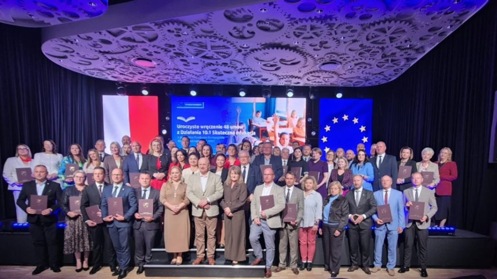 Grupa około 50 osób w formalnych ubraniach, stojących na scenie z flagą Polski, flagą Unii Europejskiej i ekranem z napisem, sufit z wzorem zębatek, fioletowe oświetlenie.