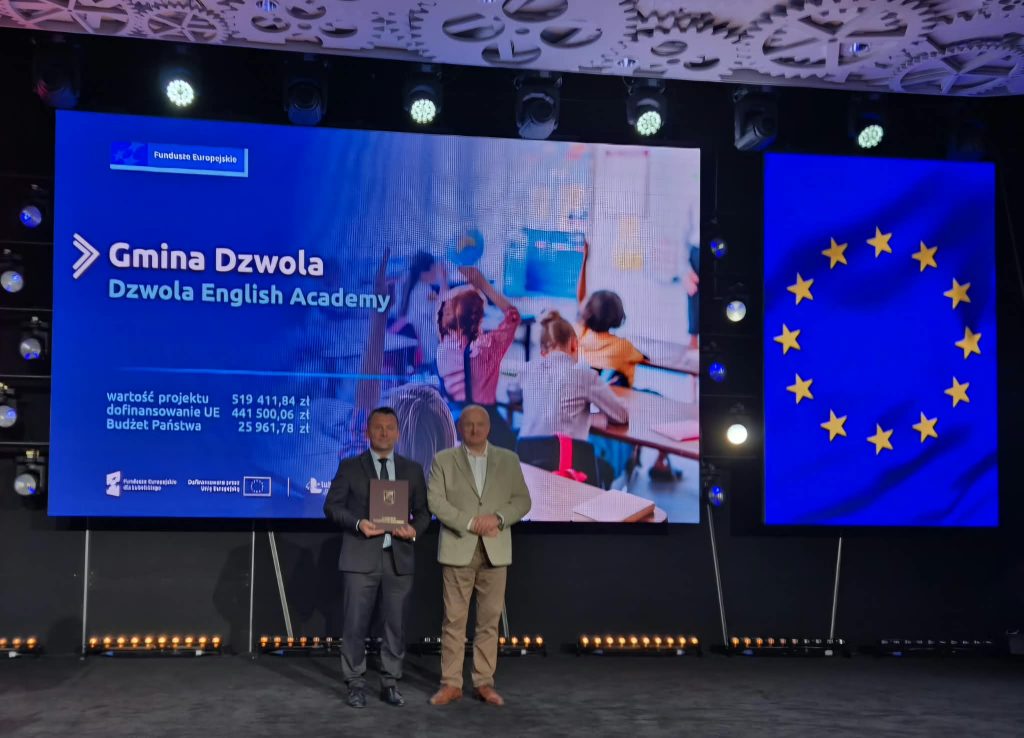 Dwóch mężczyzn na scenie na tle dużych ekranów z napisem "Gmina Dzwola Dzwola English Academy" i flagą Unii Europejskiej w kolorze niebieskim ze złotymi gwiazdami.