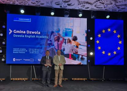 Dwóch mężczyzn na scenie na tle dużych ekranów z napisem "Gmina Dzwola Dzwola English Academy" i flagą Unii Europejskiej w kolorze niebieskim ze złotymi gwiazdami.