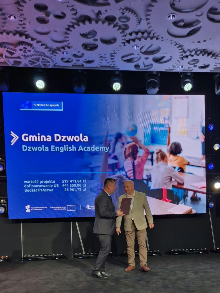 Dwóch mężczyzn na scenie na tle dużego ekranu z napisem „Gmina Dzwola Dzwola English Academy” oraz grafiką dzieci w klasie. Niebieskie i czarne barwy, sufit z dekoracją zębatek.