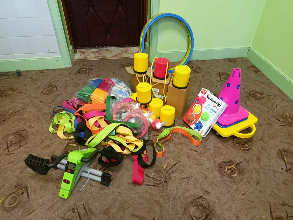 Kolorowe taśmy, hula-hop, żółte i czerwone plastikowe kubki na sznurkach, różowe pachołki, zielony rowerek treningowy na brązowym dywanie przy ścianach w jasnej zieleni.