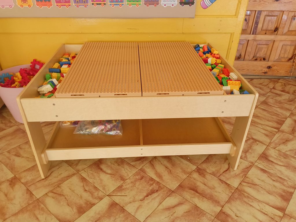 Stolik z jasnobrązową płytą do klocków, wokół kolorowe klocki w pudełku, żółta ściana i drewniane drzwi w tle, podłoga w brązowo-beżową kratę.