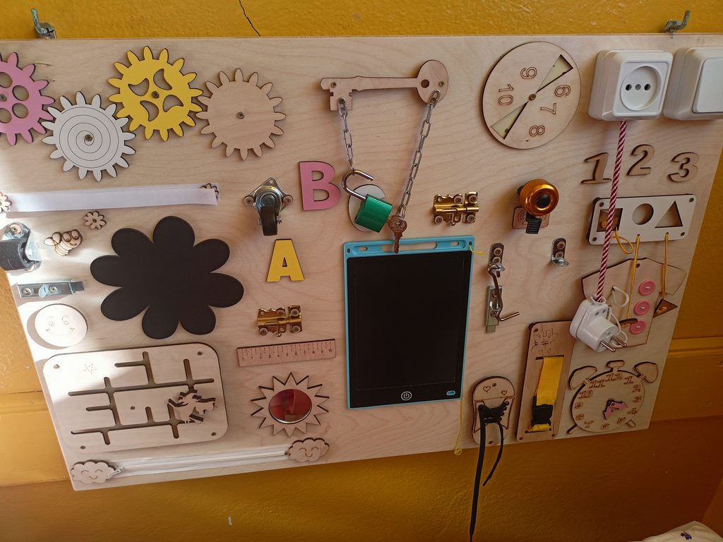 Drewniana tablica manipulacyjna z przekładniami zębatymi, kłódką i łańcuchem, kolorowymi literami A i B, liczbami 1, 2, 3, gniazdem elektrycznym, zegarem i różnymi zamkami oraz elementami do ćwiczeń manualnych.