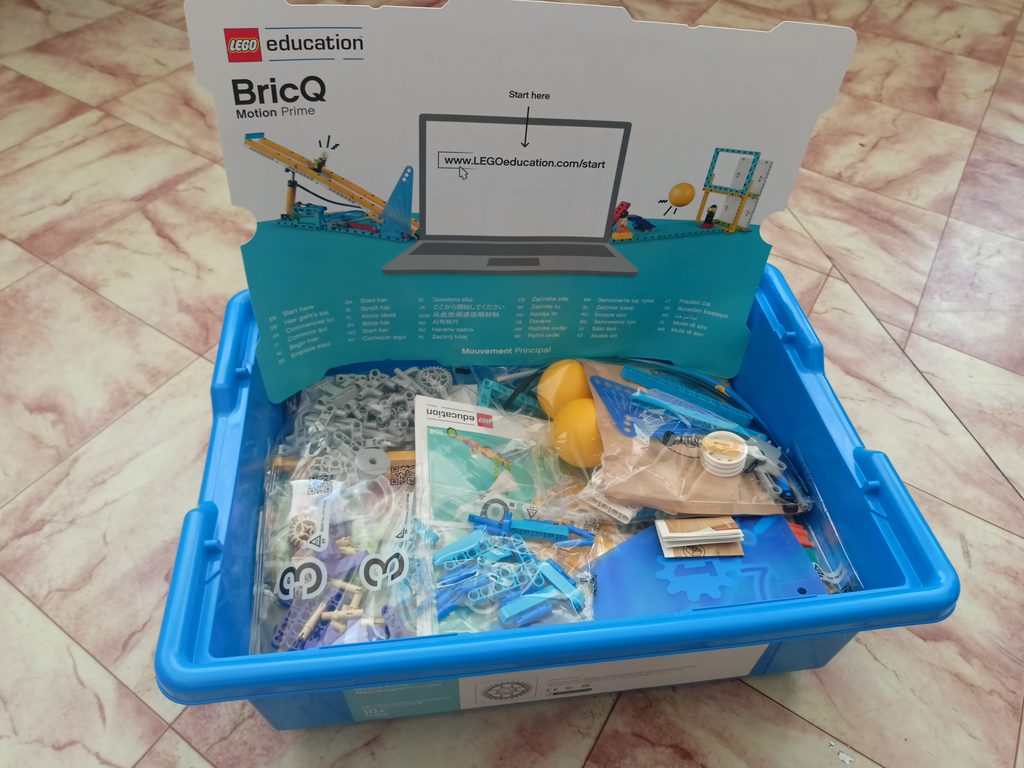 Niebieskie pudełko z elementami LEGO, instrukcje, żółte kule, szare i niebieskie plastikowe części, podkładka z nadrukiem laptopa i napisem LEGO education BricQ Motion Prime.