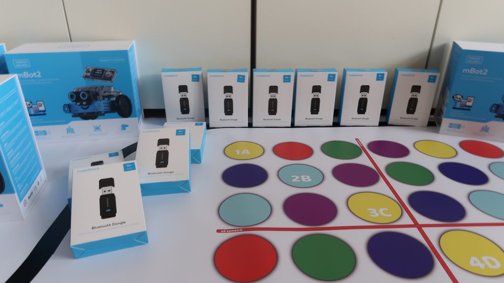 Pudełka z adapterami Bluetooth Dongle i zestawy mBot2 oraz kolorowe koła z oznaczeniami na białym tle z czerwoną linią symetrii.