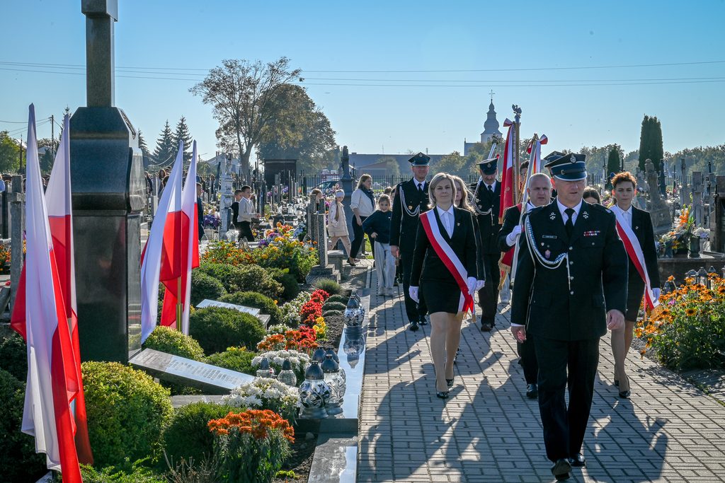Grupa ludzi w mundurach i strojach oficjalnych z biało-czerwonymi szarfami idzie brukowaną alejką na cmentarzu, po bokach groby z kolorowymi kwiatami i polskie flagi.