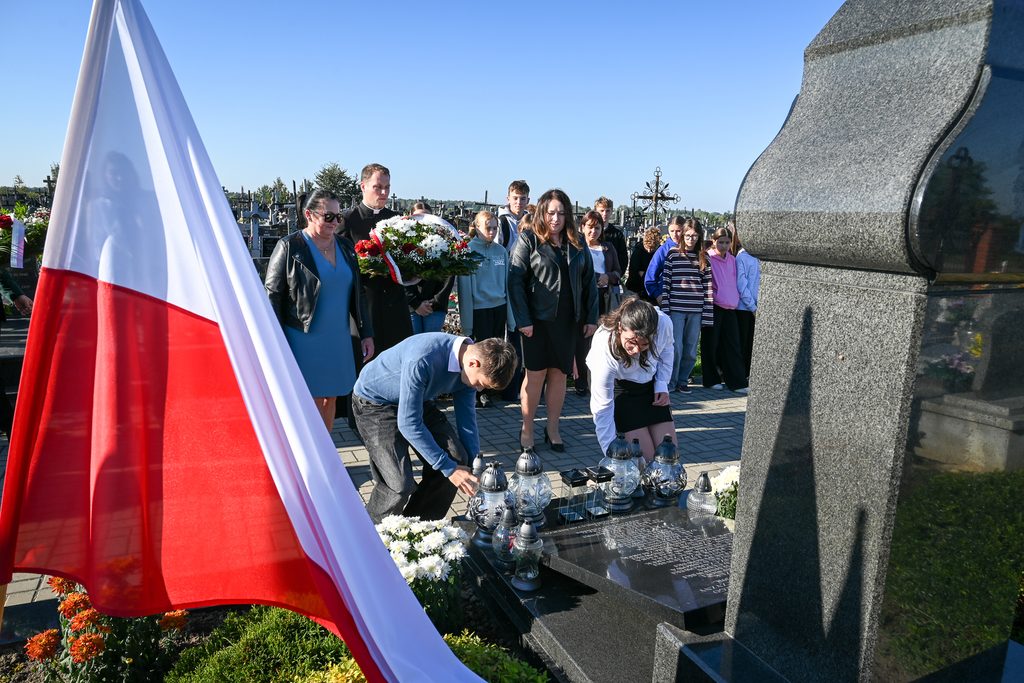 Grupa osób na cmentarzu obok nagrobka, mężczyzna w sutannie z wiązanką kwiatów, biało-czerwona flaga Polski po lewej stronie, lampiony i kwiaty na nagrobku.