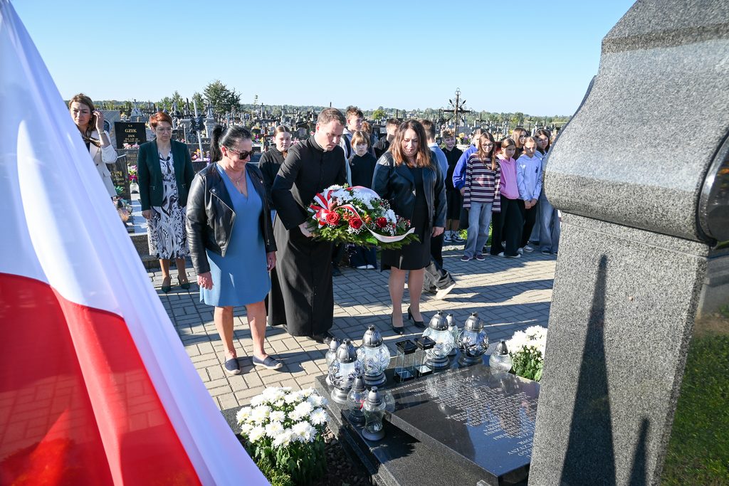 Grupa osób na cmentarzu przy nagrobku, mężczyzna w czarnym stroju niesie wieniec z czerwono-białymi kwiatami, na pierwszym planie biało-czerwona flaga.