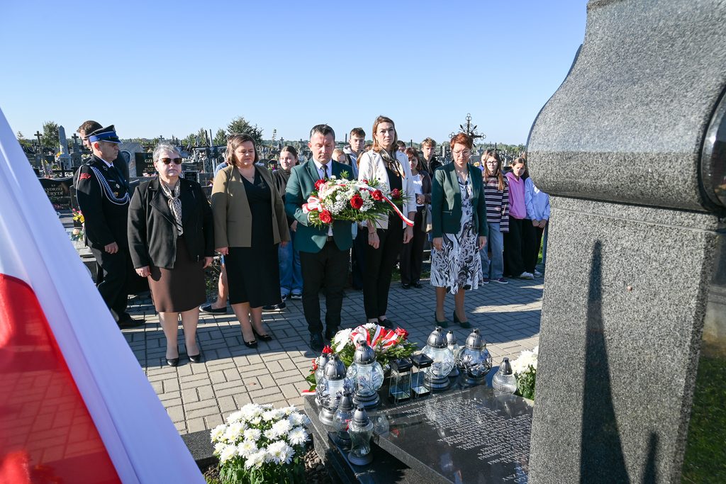 Grupa ludzi stojąca przy nagrobku na cmentarzu, mężczyzna trzyma bukiet kwiatów z czerwonymi różami, w tle liczne nagrobki i niebieskie niebo.