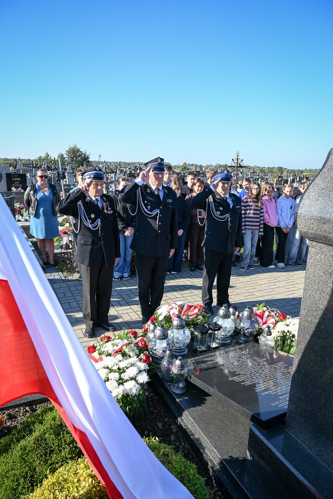 Trzech mężczyzn w mundurach strażackich oddaje honor przy pomniku na cmentarzu, obok wieńce z białych i czerwonych kwiatów oraz grupa osób w tle.