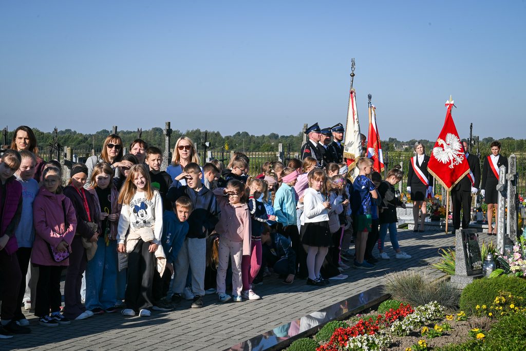 Grupa dzieci i dorosłych stoi na cmentarzu, w tle trzy osoby w mundurach z flagami i trzy osoby w strojach oficjalnych z czerwono-białymi szarfami.