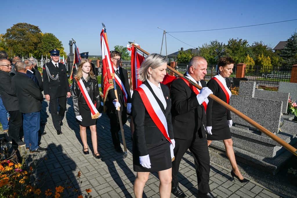Grupa osób w mundurach i strojach formalnych z biało-czerwonymi szarfami niesie flagi na cmentarzu, po prawej nagrobki z szarego kamienia, w tle zieleń i niebieskie niebo.