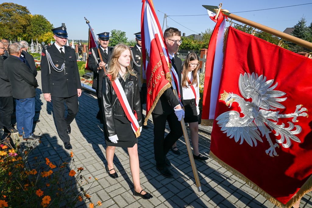 Młodzież i mężczyźni w mundurach strażackich idący po chodniku, dziewczyna w czarnej kurtce i białej szarfie, czerwone flagi z orłem białym.
