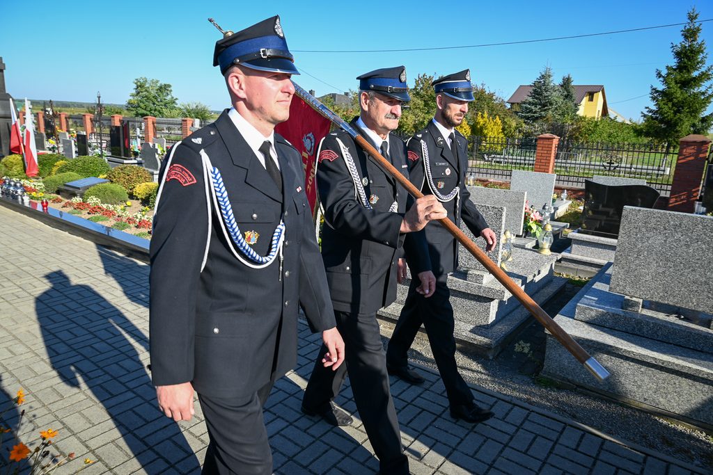 Trzech mężczyzn w mundurach strażackich z niebieskimi czapkami idzie obok nagrobków na cmentarzu, jeden niesie drzewce z flagą. Słoneczny dzień, zielone rośliny.