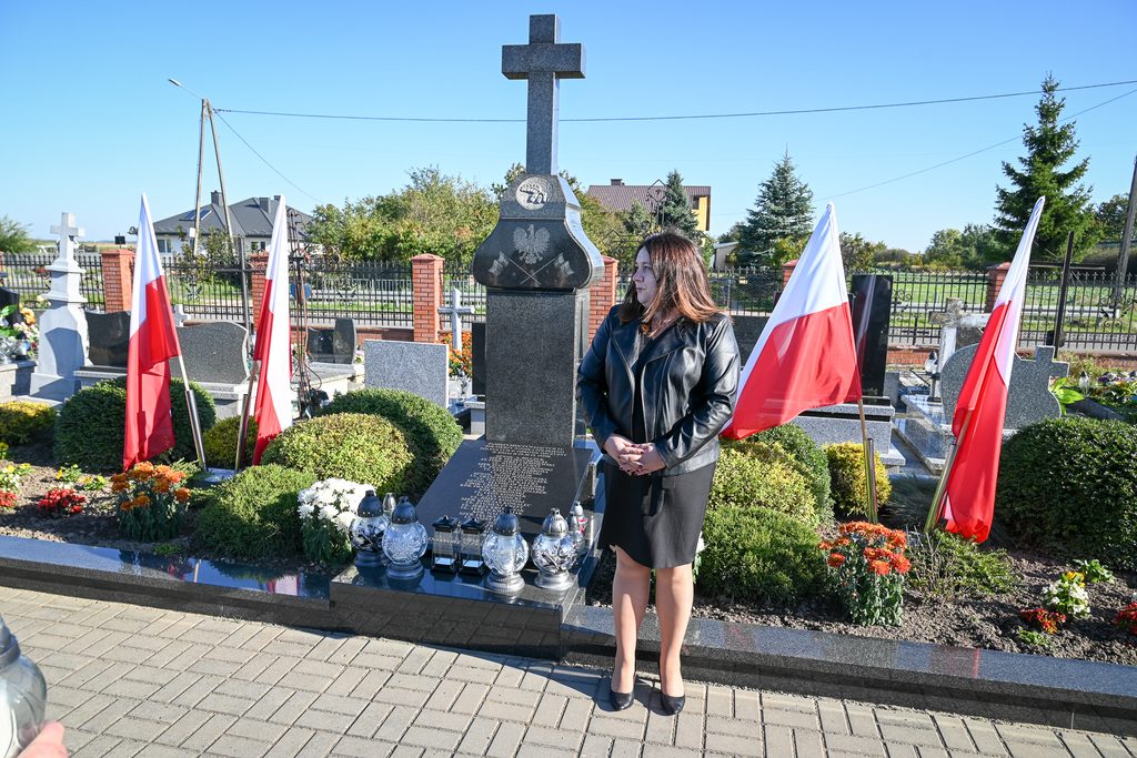 Kobieta w czarnej sukience i kurtce stoi obok czarnego nagrobka z krzyżem, otoczonego kwiatami i czterema biało-czerwonymi flagami Polski, na tle ogrodzenia i niebieskiego nieba.