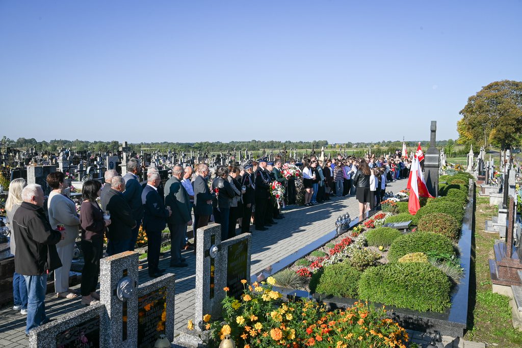 Ludzie stojący przy alei na cmentarzu, kwiaty, krzewy, polskie flagi, nagrobki, jasne niebo.