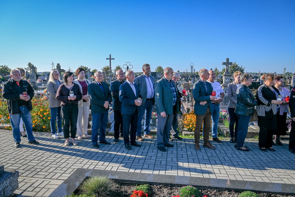 Grupa ludzi stojąca na cmentarzu na chodniku z szarych płytek, w tle liczne nagrobki i kwiaty pod jasnym, błękitnym niebem.