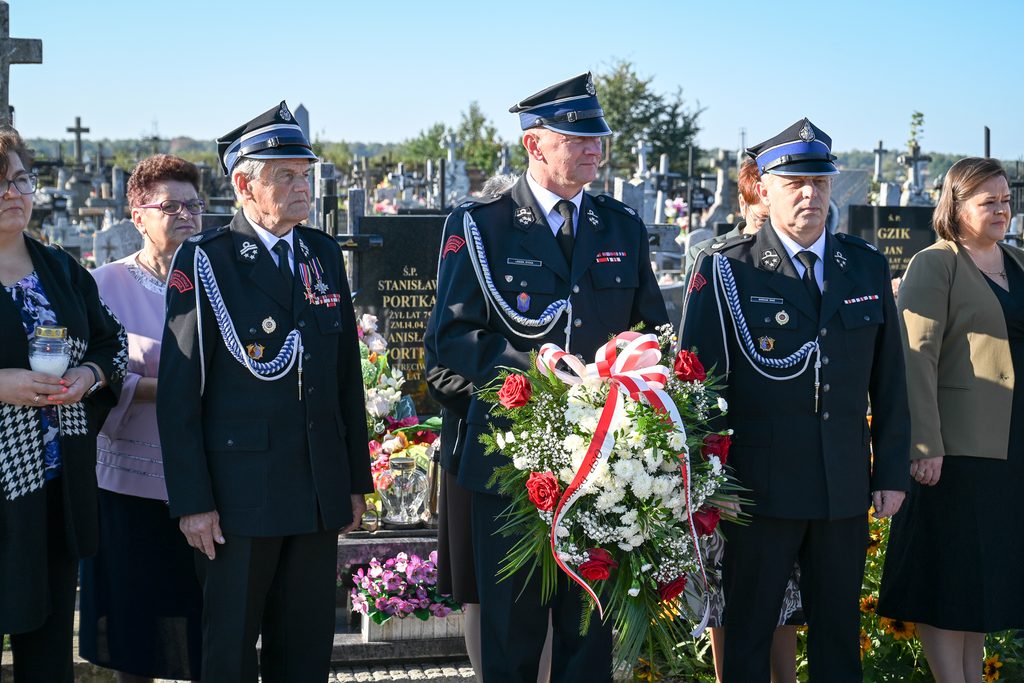 Trzech mężczyzn w mundurach strażackich z odznaczeniami i czapkami, jeden trzyma wieniec kwiatowy z czerwono-białą kokardą, w tle cmentarz i kobiety.