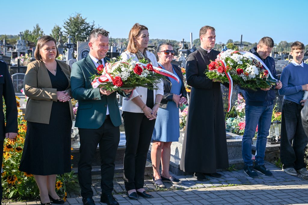 Grupa ludzi na cmentarzu, dwie osoby trzymają wiązanki kwiatów z czerwonymi różami i białymi kwiatami, w tle nagrobki i drzewa, niebieskie niebo.
