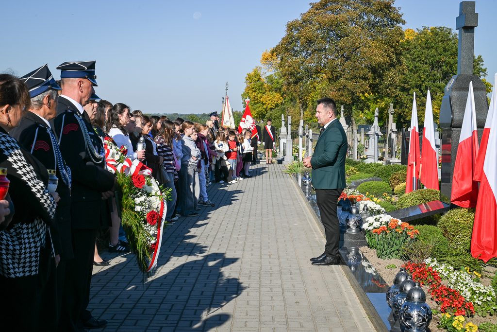 Grupa ludzi stojąca po obu stronach alejki na cmentarzu, po prawej pomnik z krzyżem i flagami Polski, kwiaty i znicze, w tle drzewa z żółto-zielonymi liśćmi.