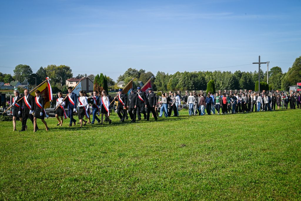 Grupa ludzi maszerujących po zielonym polu, niektórzy niosą flagi, w tle drzewa, domy i krzyż, niebo bezchmurne, słoneczny dzień.