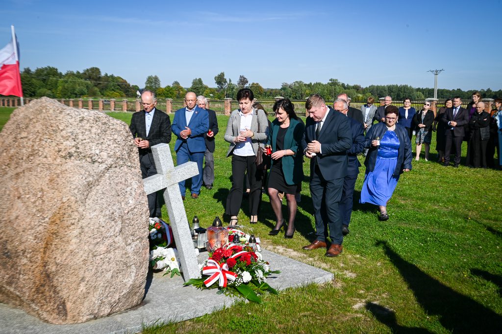 Grupa ludzi na zielonej trawie, w tle płot i drzewa, z przodu kamień, krzyż i wiązanki kwiatów z biało-czerwonymi wstążkami, kilka osób trzyma znicze.