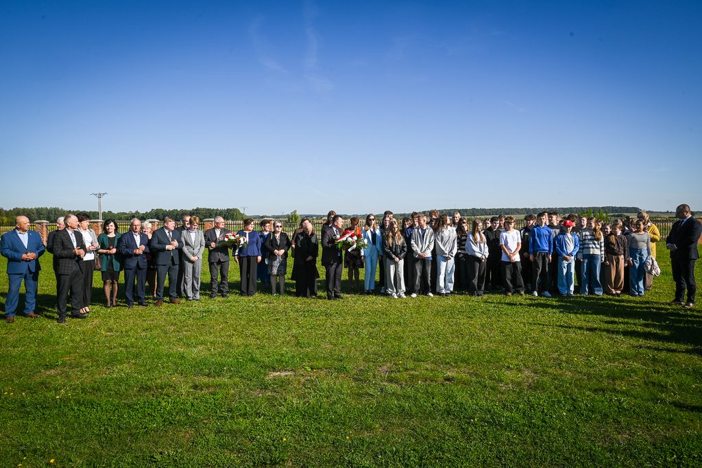 Grupa ludzi w różnym wieku stoi na zielonej trawie pod jasnym, bezchmurnym niebem, niektórzy trzymają wiązanki kwiatów, w tle niska linia drzew i ogrodzenie.
