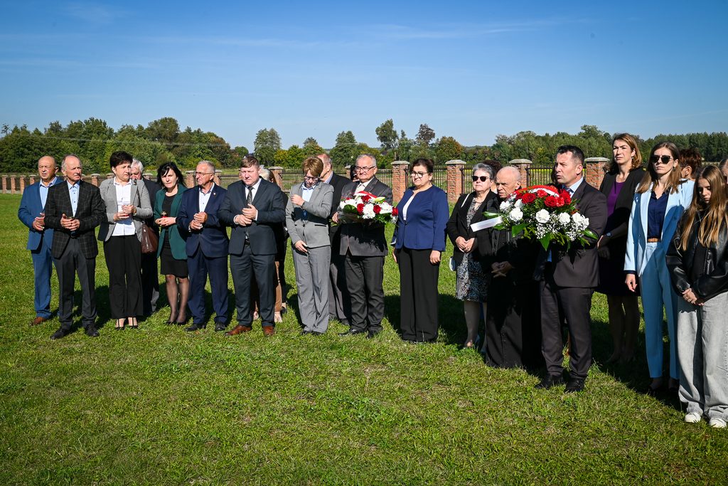 Grupa ludzi stojących na zielonej trawie w słoneczny dzień, część osób trzyma bukiety kwiatów z czerwonymi i białymi różami, w tle drzewa i mur z cegieł.