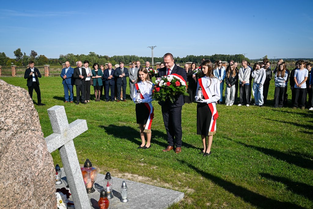 Grupa ludzi na trawie przy pomniku z kamienia i szarym krzyżem, dwie dziewczyny w białych bluzkach, czarnych spódnicach z biało-czerwonymi szarfami, mężczyzna w garniturze z wieńcem kwiatów.