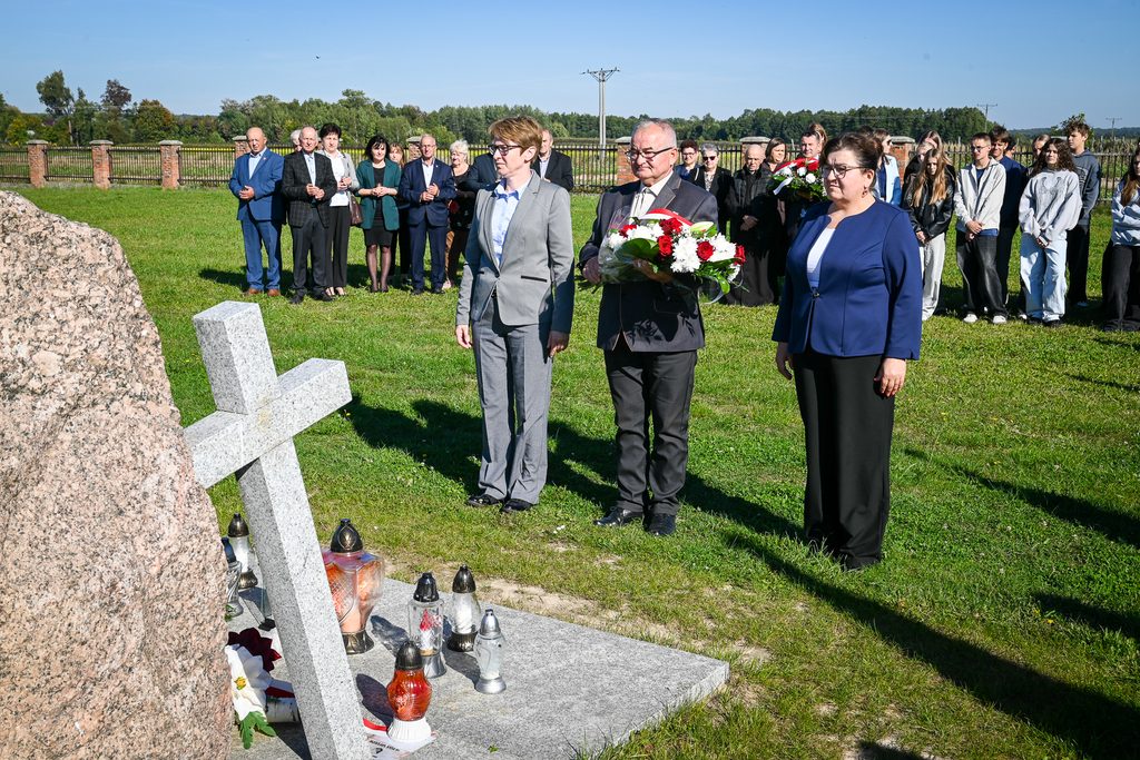 Grupa ludzi stojąca na zielonej trawie przy pomniku z jasnoszarym kamiennym krzyżem i zniczami, w tle ogrodzenie i drzewa pod błękitnym niebem.