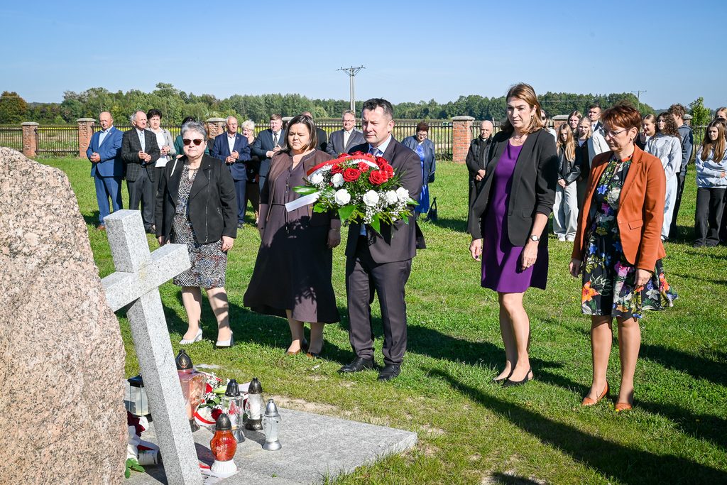 Grupa ludzi stoi na zielonej trawie przy kamieniu z szarym krzyżem i zniczami, mężczyzna trzyma bukiet czerwonych i białych kwiatów, w tle niebieskie niebo i ogrodzenie z cegieł.