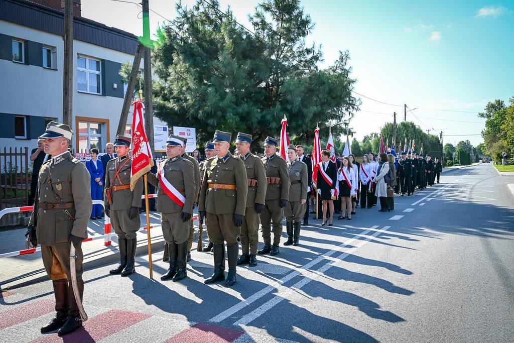 Grupa ludzi w mundurach i strojach oficjalnych stoi w szeregu na ulicy, trzymając czerwono-białe sztandary, w tle budynek i drzewa na jasnym niebie.