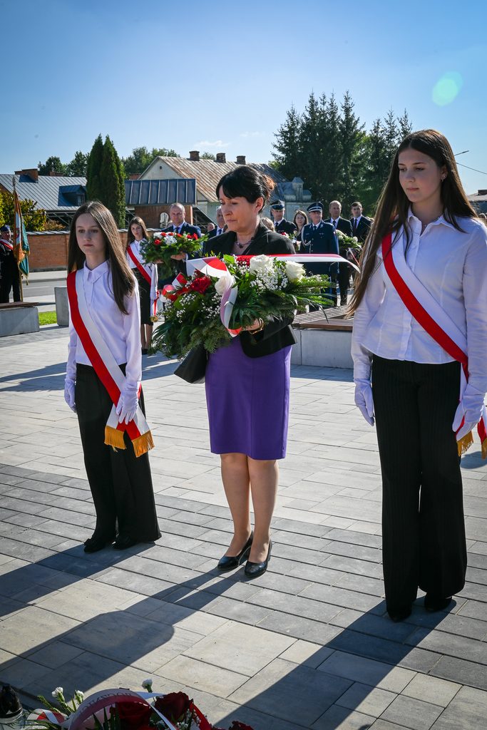 Kobieta w fioletowej spódnicy trzymająca bukiet z czerwonych i białych kwiatów, obok dwie dziewczyny w białych koszulach, czarnych spodniach i biało-czerwonych szarfach na tle ludzi i budynków.