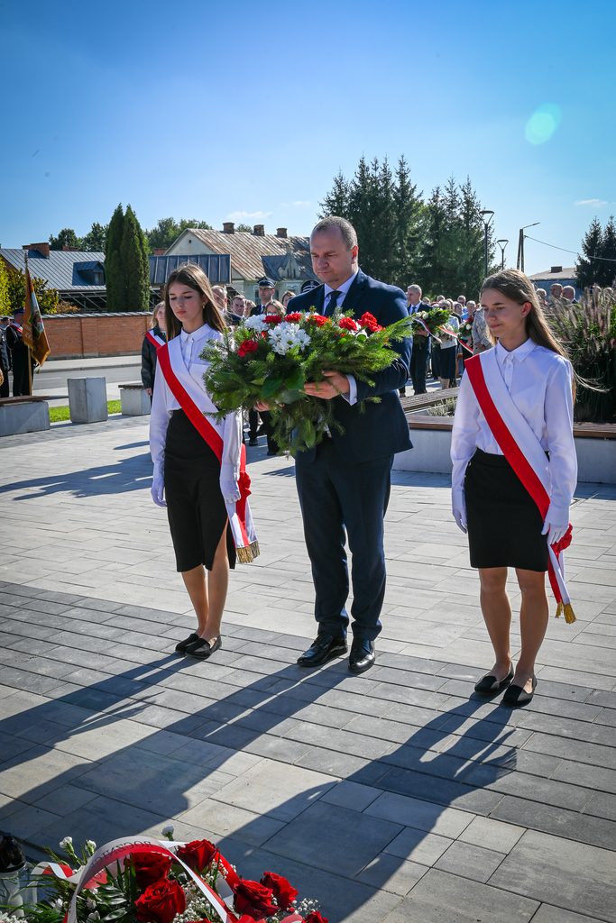 Mężczyzna w granatowym garniturze i dwie dziewczyny w białych koszulach, czarnych spódnicach i czerwono-białych szarfach niosą wieniec kwiatów na tle brukowanego placu i nieba.