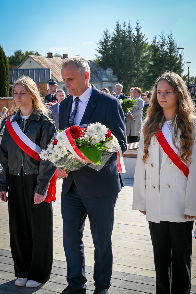 Mężczyzna w garniturze z bukietem kwiatów z biało-czerwoną wstążką oraz dwie młode kobiety w białej i czarnej odzieży z biało-czerwonymi szarfami, na tle ludzi i budynków.