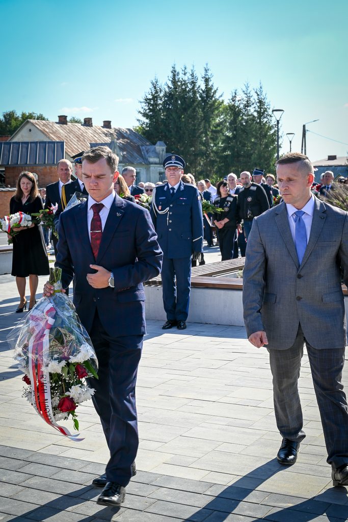 Mężczyźni w garniturach i mundurze na tle grupy ludzi, niebieskie niebo, wiązanka kwiatów z biało-czerwoną wstążką.