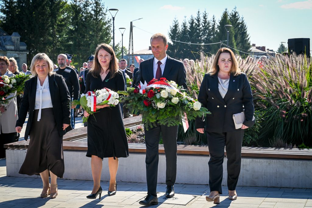Cztery osoby w formalnych strojach idące z wiązankami kwiatów, w tle grupa ludzi i wysokie trawy ozdobne, słoneczny dzień.