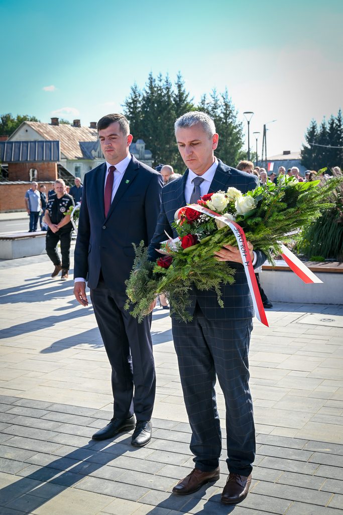 Dwóch mężczyzn w garniturach trzymających wieniec z czerwonych i białych róż oraz zielonych gałązek, na tle ludzi i budynków, w słoneczny dzień.