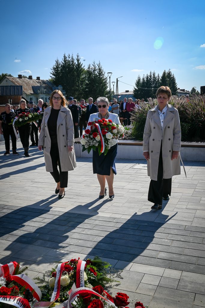 Trzy kobiety w płaszczach i sukienkach idą z wieńcami czerwonych i białych kwiatów, w tle grupa ludzi na placu z kostki brukowej pod błękitnym niebem.