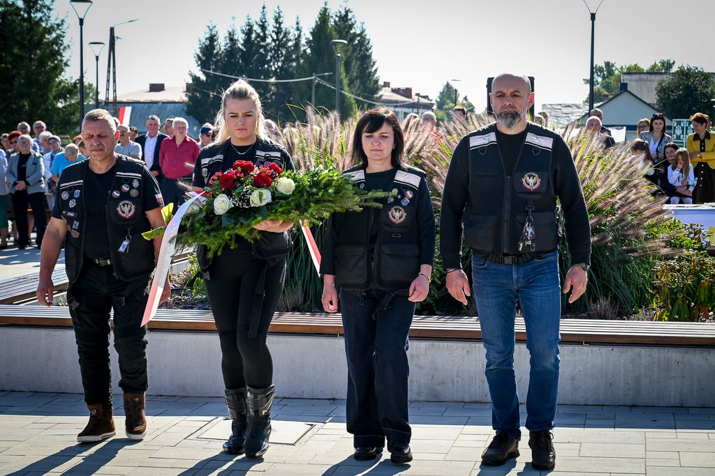 Cztery osoby z czarnymi kamizelkami z naszywkami, kobieta trzymająca bukiet czerwonych i białych kwiatów, w tle grupa ludzi, zieleń i budynki.